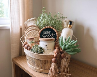 May include: A round wooden basket filled with a white candle, a bottle with a pump, a small glass jar of matches, two green succulents, a circular sign with the text Home Sweet, and a wooden bead garland with two jute tassels.