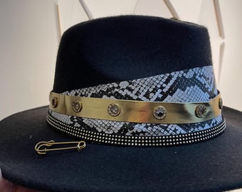 Może przedstawiać: Czarny kapelusz fedora z paskiem w wzór skóry węża, złotą ozdobą i kryształkami. Do kapelusza przyczepiona jest złota szpilka.