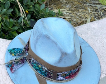 May include: A light blue fedora hat with a distressed finish. The hat features a brown leather band and a colorful patterned fabric band. A matching fabric bow is tied on the side. The hat is a stylish accessory.