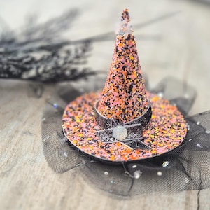 May include: A small witch's hat in pink, orange, and black glitter. It has a black ribbon and a silver decoration. The hat is set on a black tulle base, ideal for Halloween celebrations.