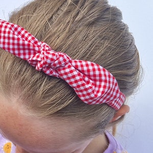 May include: A red and white gingham headband with a knotted bow. The headband is worn on a person's head.