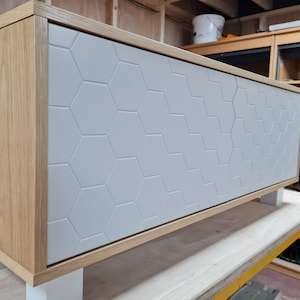 Puede incluir: Un armario de madera clara con una puerta blanca que presenta un patrón hexagonal. El armario tiene cuatro patas blancas y está sentado sobre una superficie de madera.