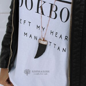 Könnte beinhalten: Ein weißes T-Shirt mit dem Text "Left My Heart In Manhattan" und einer schwarzen zahnförmigen Halskette mit Strasssteinen. Die Halskette ist an einer roségoldenen Kette.