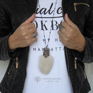 Könnte beinhalten: Eine silberne Kettenkette mit einem großen Anhänger mit einem braunen und weißen Fellbommel und einer silbernen Krone mit Strasssteinen. Der Anhänger hängt an einer silbernen Kette mit schwarzen Perlen. Die Kette wird von einer Person getragen, die ein weißes T-Shirt mit dem Text "Visual Culture Book" und eine schwarze Lederjacke trägt. Der Text "Juniper & Eloise Crafts for Life" ist auf das T-Shirt gedruckt.