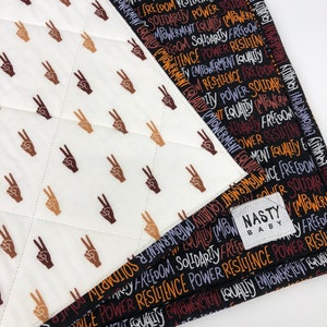 May include: A black and white baby blanket with a peace sign pattern on one side and a repeating pattern of the words "equality", "empowerment", "freedom", "solidarity", "resilience", and "power" on the other side. The blanket has a small black and white label that says "Nasty Baby".