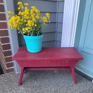 May include: A small, red wooden bench with angled legs. A turquoise flower pot filled with yellow flowers sits on top. The bench is placed outside a home with a blue door and gray siding.