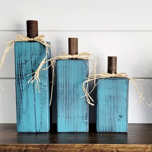 May include: Three teal blue wooden pumpkins with a rustic finish and natural raffia bows. The pumpkins are arranged in a graduated size from largest to smallest.
