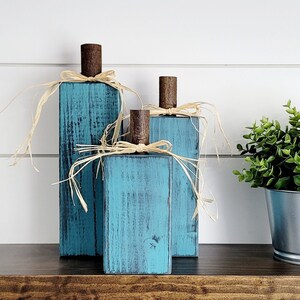 May include: Three teal blue wooden pumpkins with a rustic finish and natural raffia bows. The pumpkins are arranged in a staggered formation on a dark wood surface.