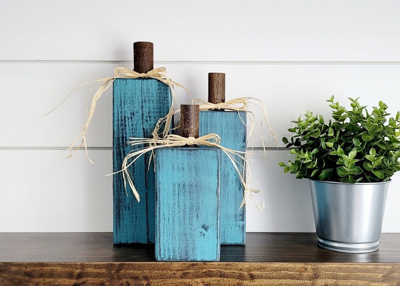 May include: Three teal blue wooden pumpkins with a rustic finish and natural raffia bows. The pumpkins are arranged in a staggered formation on a dark wood surface.