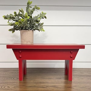 May include: A small, red wooden bench with a rectangular top and angled legs. The bench is painted a vibrant red and features two decorative rivets on each side. A potted plant with green foliage sits on top of the bench.