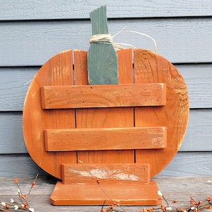 May include: A rustic, wooden pumpkin-shaped sign with an orange body and a green stem. The sign has three horizontal wooden slats and a base. The stem is tied with raffia. Perfect for fall decor.