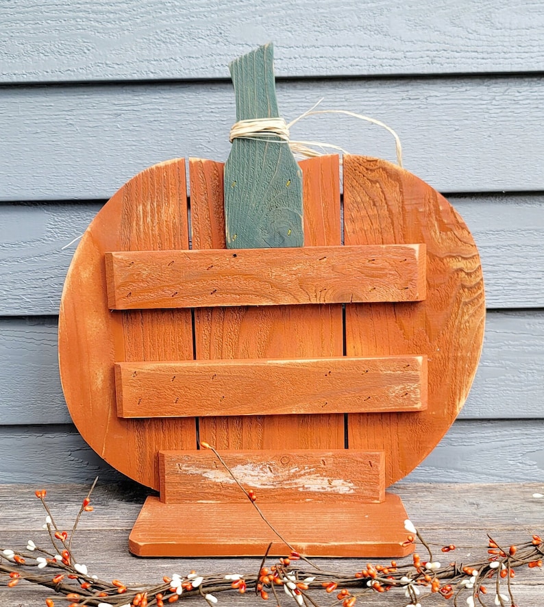 May include: A rustic, wooden pumpkin-shaped sign with an orange body and a green stem. The sign has three horizontal wooden slats and a base. The stem is tied with raffia. Perfect for fall decor.