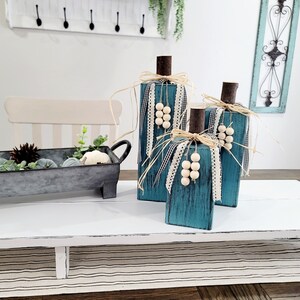 May include: Three teal blue wooden pumpkins decorated with burlap, white ribbon, and white wooden beads. The pumpkins are sitting on a white table with a white and black striped runner.
