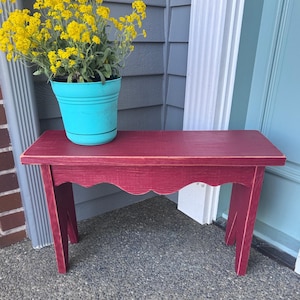 May include: A small, burgundy-colored wooden bench with a scalloped edge. A turquoise flower pot with yellow flowers sits on top. The bench has four angled legs and is placed on a gray surface.