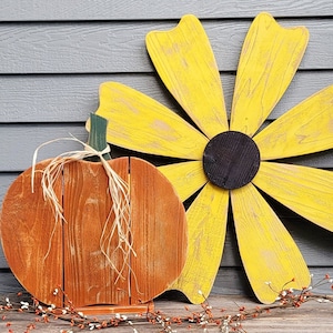 May include: Wooden fall-themed decorations featuring a pumpkin and a large flower. The pumpkin is orange with a raffia stem, and the flower has yellow petals and a black center. The decorations are set against a gray background.