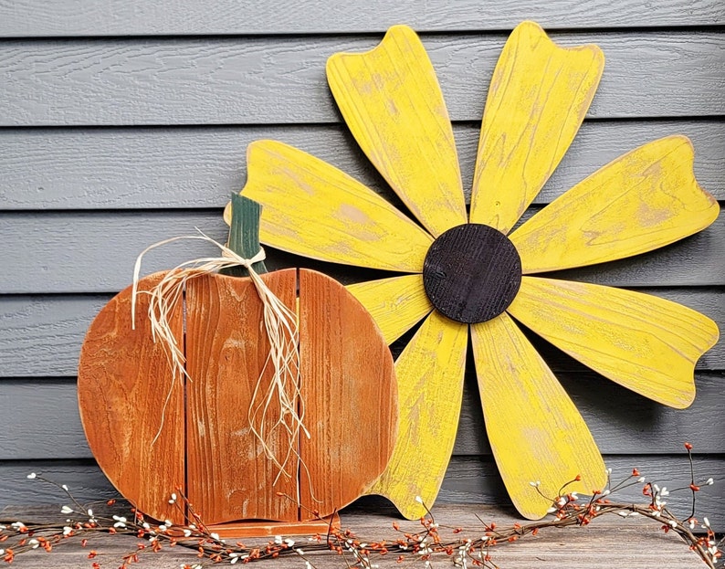 May include: Wooden fall-themed decorations featuring a pumpkin and a large flower. The pumpkin is orange with a raffia stem, and the flower has yellow petals and a black center. The decorations are set against a gray background.