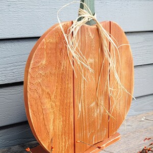 May include: A wooden pumpkin decoration with an orange body and a green stem. The pumpkin is made of vertical wooden planks and sits on a small base. Natural raffia is tied around the stem. A fall-themed decorative item.
