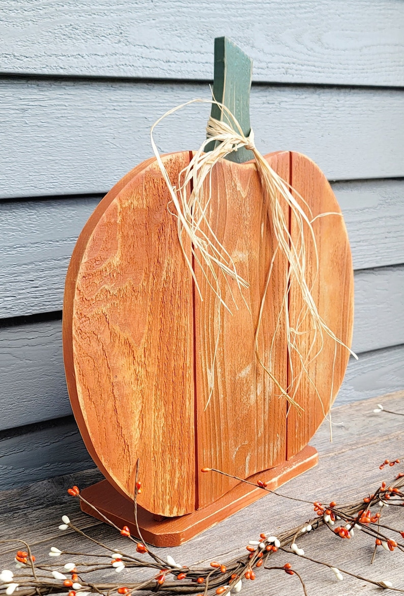 May include: A wooden pumpkin decoration with an orange body and a green stem. The pumpkin is made of vertical wooden planks and sits on a small base. Natural raffia is tied around the stem. A fall-themed decorative item.