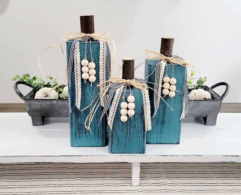 May include: Three teal blue wooden pumpkins decorated with white beads, black and white checkered ribbon, and lace ribbon. The pumpkins are sitting on a white table with a black and white striped runner.