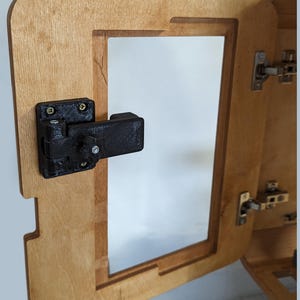 Peut inclure: Un meuble en bois partiellement ouvert avec un loquet noir. Le meuble est en bois de couleur claire et possède une ouverture rectangulaire. Le loquet noir est fixé à la porte et au cadre du meuble.
