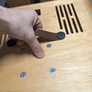 Puede incluir: Un panel de madera de color marrón claro con un asa de correa de cuero marrón asegurada con cierres circulares negros. La superficie incluye una serie de ranuras de ventilación verticales y cuatro tornillos metálicos empotrados en el.