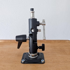 Op de afbeelding: Een zwarte fotografie lichtstandaard met een zilveren instelknop en een zwarte basis met een zilveren schroef. De standaard staat op een houten oppervlak.