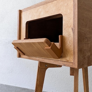 Puede incluir: Una mesa auxiliar de madera con un estante inclinado único y un pequeño compartimento abierto. La mesa está hecha de madera contrachapada y tiene cuatro patas.
