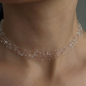 May include: A clear, multi-strand necklace with small, round beads. The necklace is worn around the neck and appears delicate and lightweight. The beads are evenly spaced along the strands, creating a subtle, sparkling effect.