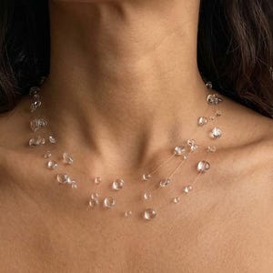 May include: A delicate necklace featuring clear, round beads of varying sizes strung on nearly invisible strands. The beads appear to float around the neck, creating a minimalist and elegant look. The necklace is a simple yet striking accessory.