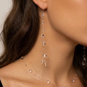 May include: Elegant silver earrings with clear, spherical beads suspended on delicate strands. A matching necklace with similar beads complements the earrings. The jewelry is displayed against a neutral background, highlighting the sparkling design.
