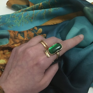 May include: A gold ring with a large, oval-shaped green gemstone. The ring is worn on the index finger of a hand.