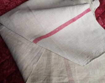 May include: Folded beige linen towel with a diagonal red stripe. The towel has a textured weave and a simple design. The background is a dark red, patterned fabric.