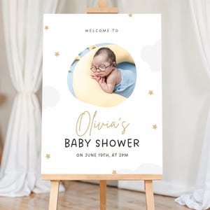 K&ouml;nnte beinhalten: Willkommen zur Baby Shower von Olivia Schild mit wei&szlig;em Hintergrund, blauen und wei&szlig;en Wolken, goldenen Sternen und einem Foto eines Babys mit Brille, das auf einem gelben Halbmond schl&auml;ft.
