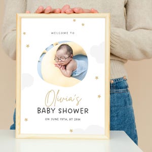 K&ouml;nnte beinhalten: Eine Babyparty-Einladung mit wei&szlig;em Hintergrund und goldenen Sternen. Die Einladung zeigt ein Foto eines Babys, das auf einem gelben Halbmond schl&auml;ft. Der Text lautet "Willkommen zur Babyparty von Olivia am 19. Juni um 14:00 Uhr."