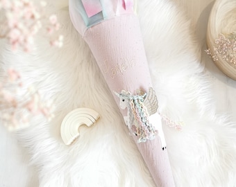 May include: A pink and white unicorn school cone with the name "Javiella" embroidered on the side. The cone is decorated with a rainbow and glitter.