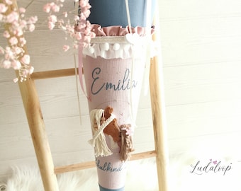 May include: A pink and blue school cone with a horse design and the name "Emilia" embroidered on it. The cone is decorated with white lace and a white bow. The text "Schulkind" is also visible on the cone.