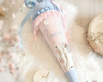 May include: A cone-shaped fabric gift bag in pastel pink and light blue, featuring a unicorn appliqué with silver wings and yarn mane. The name Lorena is embroidered in blue script. It is topped with a light blue fabric closure and satin ribbons.