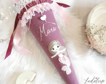 May include: A pink and white school cone with a mermaid design and the name "Mara" embroidered on it. The cone is decorated with white glitter and a pink ribbon.
