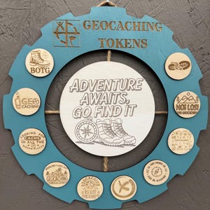 May include: A blue wooden gear-shaped sign with the words "GEOCACHING TOKENS" engraved on it. The sign has a white center with the text "ADVENTURE AWAITS, GO FIND IT" and several wooden tokens with various designs and text related to geocaching.