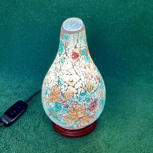 May include: A mosaic glass lamp in a teardrop shape. The lamp has a colourful mosaic pattern with white, blue, orange, and red hues. It rests on a dark brown wooden base and has a black power cord with an on/off switch.