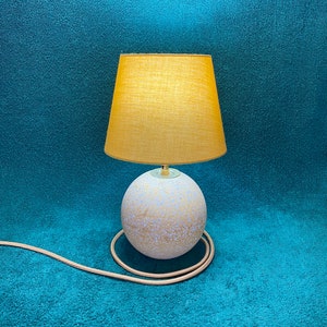 May include: A decorative table lamp with a spherical base and a mustard-yellow lampshade. The base is speckled with blue and tan, and the cord is a matching tan colour. The lamp is designed for indoor use.