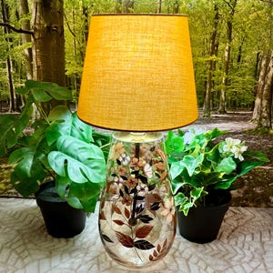May include: A table lamp with a tapered mustard yellow fabric lampshade and a clear glass base. The glass base features a painted botanical design with brown and white flowers and dark brown leaves on a slender central stem inside the glass.