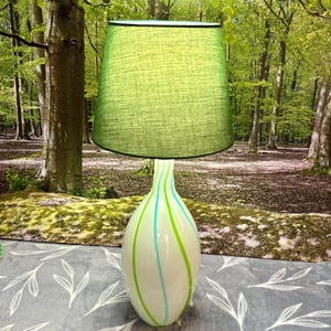 May include: A table lamp with a green lampshade and a white base. The base has vertical stripes in green and blue. The lamp is set against a backdrop of a forest. The lamp is a decorative item.