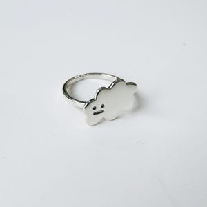 May include: Silver ring featuring a cloud-shaped design with a simple face drawn on it.