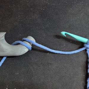 May include: A close-up of crochet tools and yarn. A gray yarn holder is connected to a blue yarn strand. A turquoise crochet hook is connected to a chain of blue yarn. The background is a dark gray surface.