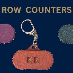 以下が含まれることがあります： 3つの行カウンター。それぞれ色が異なり、数字が異なります。カウンターは銀色のキーチェーンに取り付けられています。カウンターは赤、紫、グレーです。カウンターの数字は87、27、13です。