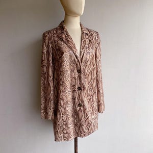 Puede incluir: Un blazer de manga larga con estampado de piel de serpiente en tonos marrón y beige. El blazer tiene cuello con solapas, tres botones y se exhibe en un maniquí. La prenda es de estilo vintage o retro.