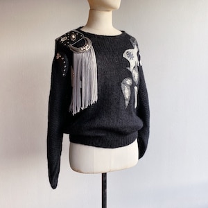 Peut inclure: Pull en maille noire avec un détail de frange argentée et noire sur l'épaule et un motif d'applique en cuir blanc et noir sur le devant.