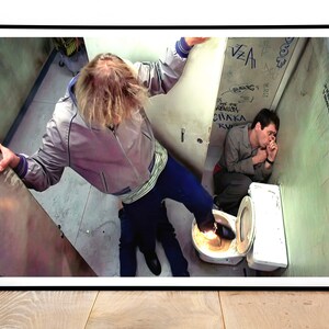 K&ouml;nnte beinhalten: Ein Schwarzwei&szlig;foto von zwei M&auml;nnern in einem Badezimmer. Ein Mann steht auf einer Toilette mit seinem Fu&szlig; auf dem Kopf des anderen Mannes. Der Mann auf der Toilette tr&auml;gt eine graue Jacke und eine blaue Jeans. Der Mann auf dem Boden tr&auml;gt ein graues Hemd und eine blaue Jeans. Die Badezimmerw&auml;nde sind mit Graffiti bedeckt.