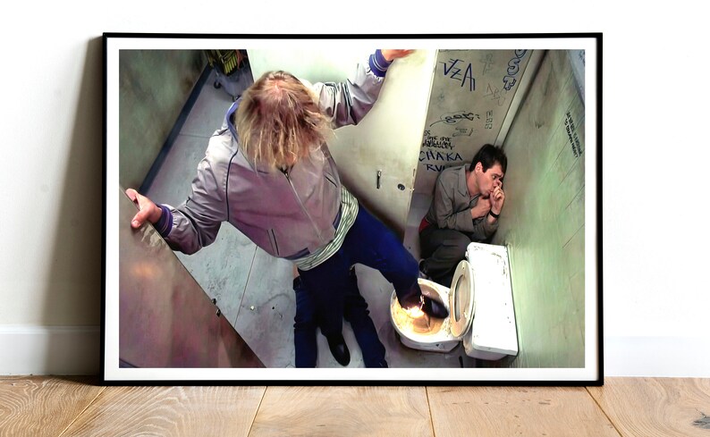 K&ouml;nnte beinhalten: Ein Schwarzwei&szlig;foto von zwei M&auml;nnern in einem Badezimmer. Ein Mann steht auf einer Toilette mit seinem Fu&szlig; auf dem Kopf des anderen Mannes. Der Mann auf der Toilette tr&auml;gt eine graue Jacke und eine blaue Jeans. Der Mann auf dem Boden tr&auml;gt ein graues Hemd und eine blaue Jeans. Die Badezimmerw&auml;nde sind mit Graffiti bedeckt.